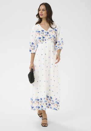 Kobieta idąca, ubrana w białą maxi sukienkę w niebieskie kwiaty, czarne sandały na paski i niosąca małą czarną plecioną kopertówkę.