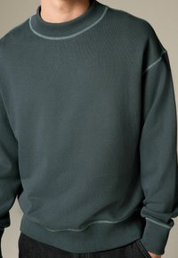 Tmavo zelená mikina s rebrovaným golierom a manžetami. Obsahuje viditeľné švy a voľný strih, vyrobená z hladkej, textúrovanej tkaniny.