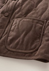 Braunes Cordstoffmaterial mit sichtbaren Nähten, das eine quadratische Tasche und einen mit Stoff bezogenen Knopf an einem gesteppten Bekleidungsstück zeigt.