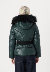 Padded green jacket with a black faux fur hood and a black belt at the waist. Features a shiny texture and chevron pattern on the back.