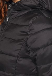 Svart puffjacka med quiltat mönster, huva och dragkedjestängning. Slät, glänsande textur med subtil glans och avslappnad passform.