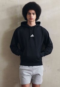 Črna kapuca z belim Adidasovim logotipom, ki ima sprednji žep, kapuco na vrvico in rebraste manšete, v kombinaciji s sivo kratkimi hlačami.