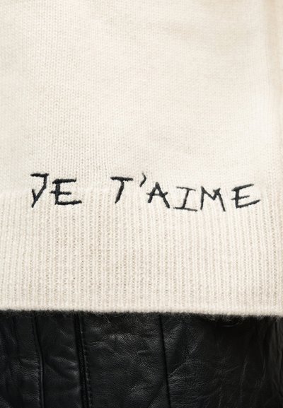 Κρεμ πλεκτό ύφασμα με μαύρη κεντητική που γράφει «JE T'AIME» πάνω από μαύρο υλικό από υφή δέρματος.