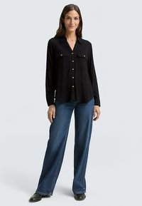 Camicia nera a maniche lunghe con colletto, due tasche sul petto e chiusura con bottoni; abbinata a jeans blu a gamba larga e scarpe nere.