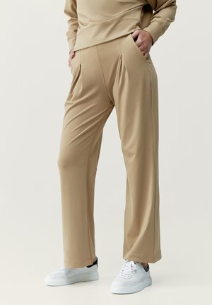 Pantaloni beige a gamba larga in tessuto liscio, con pieghe frontali, tasche laterali e vestibilità rilassata, abbinati a scarpe da ginnastica bianche.