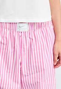 Pajama kratke hlače s črtami v roza barvi, izdelane iz gladkega materiala, z elastičnim pasom in belim oznako Nike na sprednji strani.