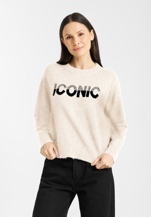Crème, fluffy trui met een ronde halslijn en zwarte "ICONIC" tekst. Gepaard met zwarte broeken, die een losse, relaxte pasvorm tonen.