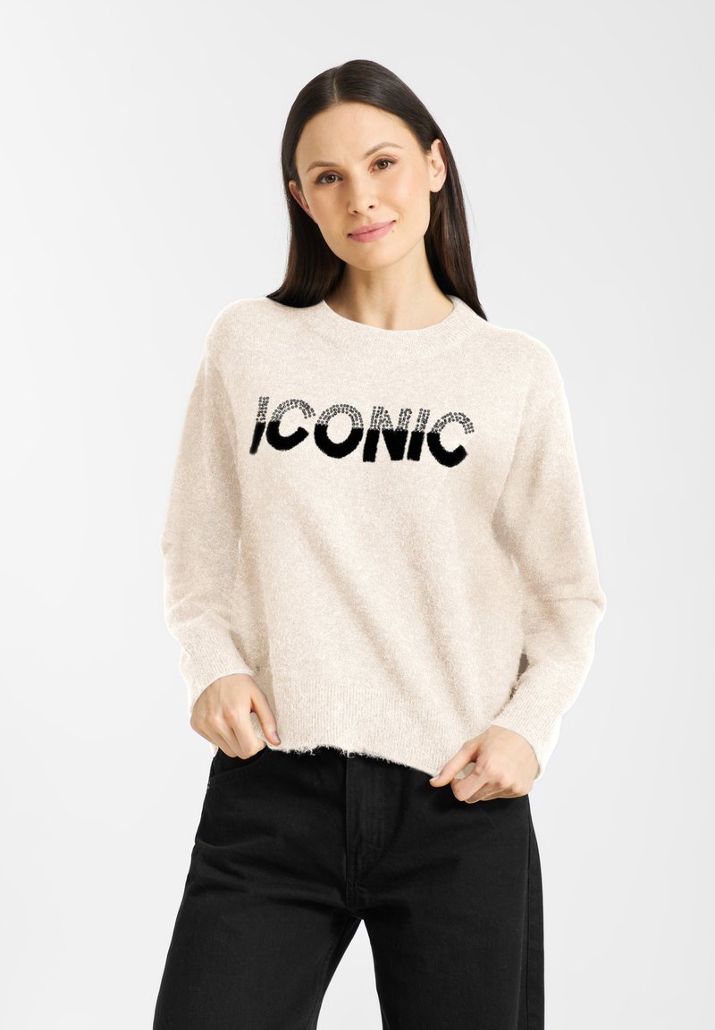 Kremowy, puszysty sweter z okrągłym dekoltem i czarnym napisem "ICONIC". Połączony z czarnymi spodniami, które mają luźny, swobodny krój.