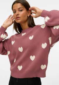 Roze trui met een wit hartpatroon, zachte textuur, ronde hals, oversized pasvorm en geribbelde manchetten, gestyled met zwarte broeken.