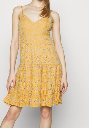 Femme portant une robe jaune sans manches avec des motifs floraux en œillets blancs et un ourlet à volants, debout devant un fond clair uni.