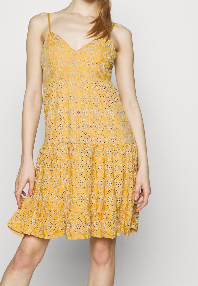 Femme portant une robe jaune sans manches avec des motifs floraux en œillets blancs et un ourlet à volants, debout devant un fond clair uni.