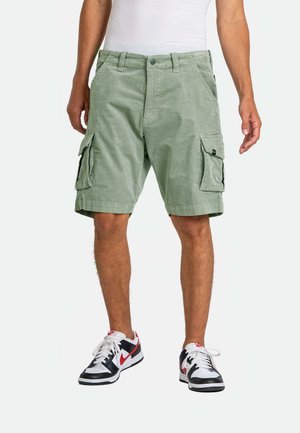 Man wearing light green cargo shorts and black, white, and red sneakers, standing with hands relaxed at sides.