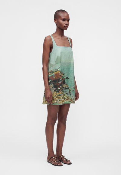 Femme mince portant une robe sans manches à encolure carrée avec un imprimé coloré de ville côtière, associée à des sandales plates noires avec des accents dorés décoratifs.