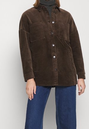 Kvinde iført en brun fløjlsjakke med knapper over en mørk rullekrave og blå jeans, stående mod en ensfarvet baggrund.