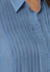 Camicia azzurra chiaro con bottoni, caratterizzata da pieghe verticali, colletto classico e piccoli bottoni coordinati per la chiusura. Tessuto morbido e strutturato.