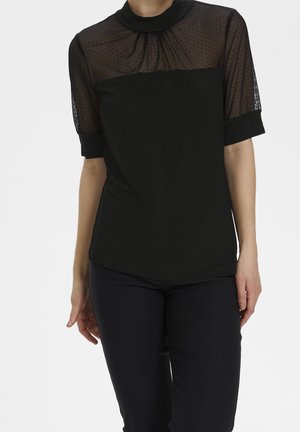 Kobieta ubrana w czarną bluzkę z przezroczystymi, kropkowanymi, krótkimi rękawami oraz dopasowane czarne spodnie, stojąca na jednolitym tle.