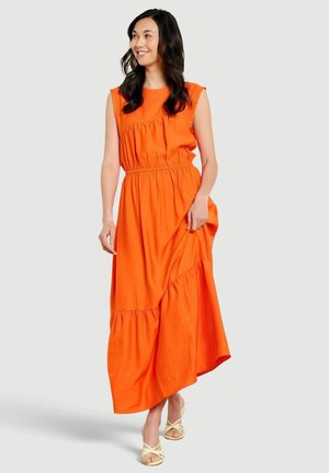 Lachende vrouw met lang donker haar staat, draagt een mouwloze oranje maxi-jurk en crème kleurige sandaaltjes met bandjes, houdt de zoom van de jurk aan één zijde vast.