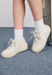 Beige sneakers with light purple accents, smooth leather upper, round toe, lace-up closure, and textured rubber sole. Worn with white crew socks.