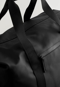 Black duffel bag with a smooth, matte texture, featuring sturdy black nylon straps and a zippered pocket. Simple and functional design.