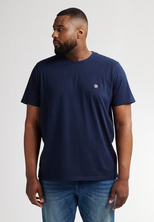 Camiseta de algodón azul marino con escote redondo, mangas cortas y un parche de logo en el lado izquierdo del pecho. Combinada con jeans de color azul claro.