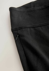 Fekete sportos leggings sima textúrával, széles derékpánttal és egy kis oldalsó cipzáras zsebbel. Látszanak a varrások részletei.