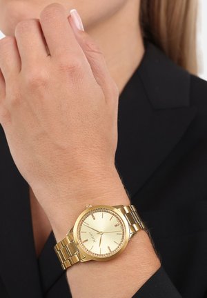 Goldene Armbanduhr mit rundem Gehäuse und passendem Metallarmband; verfügt über ein helles Zifferblatt mit Stundenmarkierungen und einem Strassbord.