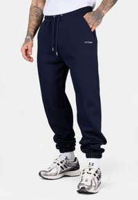 Navyblaue Sweatpants mit einem Kordelzugbund, Bündchen an den Knöcheln und gesticktem Logo auf dem Oberschenkel, kombiniert mit silbernen Laufschuhen.