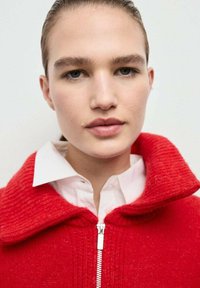 Giacca rossa lavorata a maglia con un ampio colletto, chiusura con zip argentata, indossata sopra una camicia bianca con colletto. La texture è a coste e morbida.