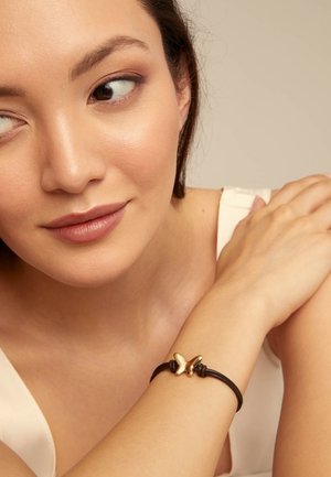Jeune femme à la peau lisse portant un bracelet en corde noire avec un pendentif papillon en or au poignet, reposant son bras sur son épaule.
