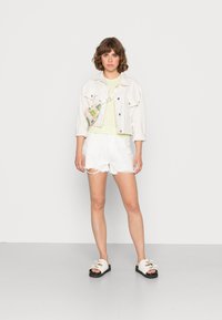Jaqueta de ganga branca sobre uma blusa xadrez em tons pastel, combinada com calções brancos desfiados e sandálias bege com solas pretas. Mala colorida com padrões usada na transversal.