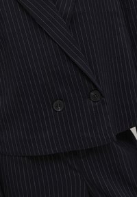 Blazer noir croisé à fines rayures avec deux boutons noirs, montrant le revers et le tissu rayé assorti.