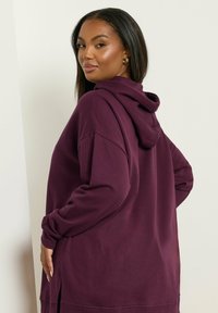 Sudadera de gran tamaño en mezcla de algodón color burdeos oscuro, con un bolsillo frontal tipo canguro, aberturas laterales y capucha con cordón. Textura suave.