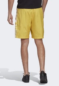 Gula badbyxor med sidofickor, elastisk midja och vita Adidas-ränder på benen. Skor i svart, står sett bakifrån.