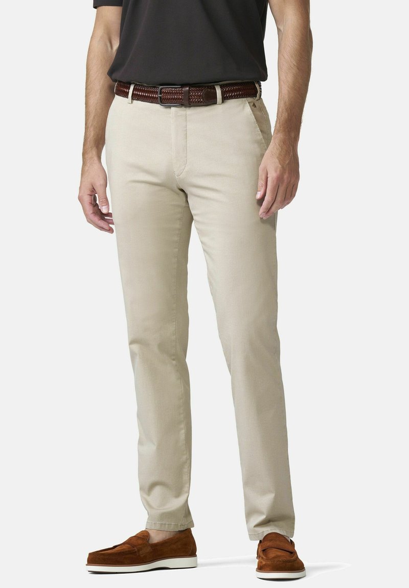 Man met beige chino's en een bruine gevlochten riem, een zwart shirt en bruine suède loafers staat tegen een effen achtergrond.