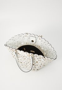 Bolso tote cortado en blanco con borde en forma de ola, cuenta con hardware dorado y un interior negro. Las asas están unidas con correas de cuero.