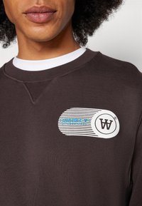 Brauner Sweatshirt mit einem Logo-Patch mit blauen und weißen Streifen und dem Text "DOUBLE V by WOOD WOOD" im linken Brustbereich.