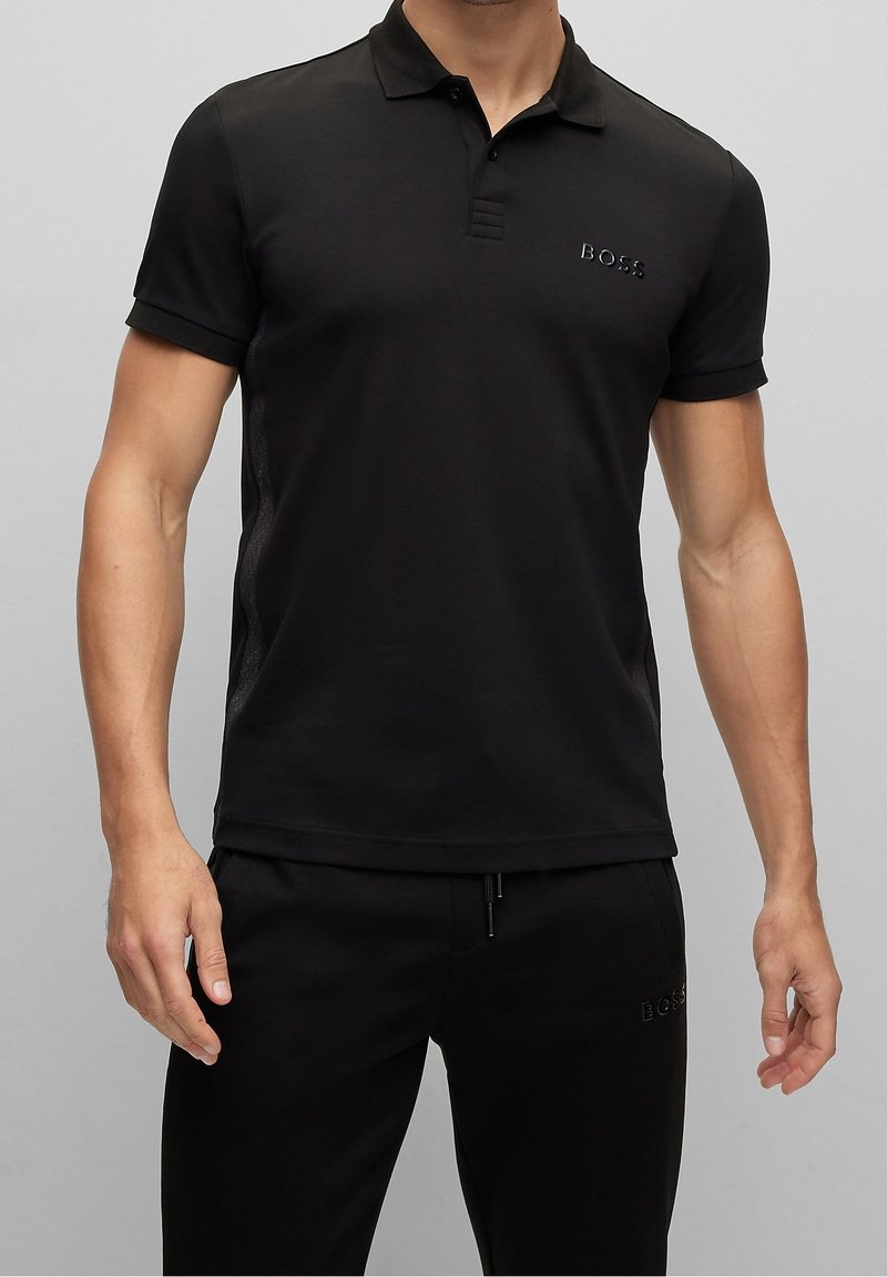 Polo noir en tissu lisse avec manches courtes, patte de boutonnage à trois boutons et détails en mesh sur les côtés. Présente un logo en relief subtil.