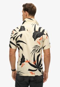 Camisa de manga corta con cuello en color crema, con un patrón de hojas tropicales en negro y naranja. Tejido suave, corte relajado, con un detalle en el yugo trasero.