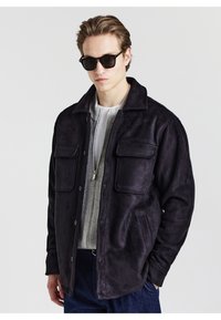 Schwarze Wildlederjacke mit zwei Fronttaschen und einem klassischen Kragen, über einem grauen Zip-Pullover getragen, kombiniert mit dunklen Sonnenbrillen.