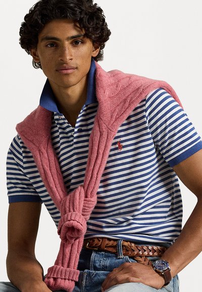 Pasiasta niebiesko-biała koszulka polo z różowym swetrem dzierganym związanym na ramionach. Noszona z dżinsami i plecionym brązowym paskiem.