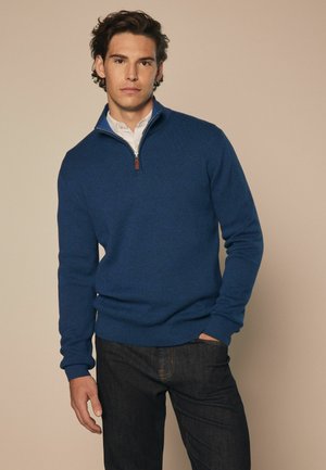 Blue knitted sweater with a zippered collar, paired with dark jeans. The sweater features a smooth texture and ribbed cuffs and hem.
