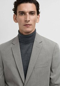 Blazer gris clair sur col roulé gris foncé, avec une texture lisse, une coupe ajustée et une poche poitrine unique. Design minimaliste.