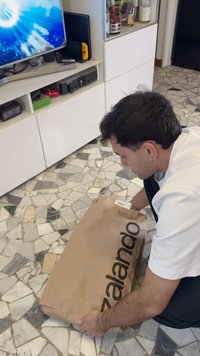 Smeđi papirnati paket označen logom "zalando", koji drži osoba na podu s uzorkom od kamena. U pozadini se nalaze TV i polica.