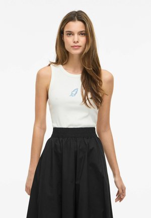 Jeune femme aux longs cheveux portant un haut blanc sans manches avec un motif coquillage bleu et une jupe noire taille haute, fond neutre.