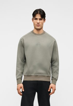 Νέος άνδρας που φοράει ελιάς πράσινη μπλούζα με στρογγυλή λαιμόκοψη και μαύρο παντελόνι, στέκεται απέναντι σε απλό ανοιχτό γκρι φόντο, κοιτάζοντας μπροστά.