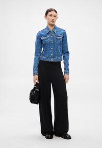 Modrá rifľová bunda s gombíkovým zapínaním a náprsnými vreckami, skombinovaná s čiernymi širokými nohavicami a malou čiernou kabelkou.