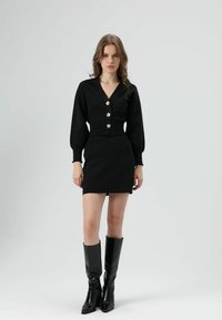 Zwarte gebreide top met lange pofmouwen, V-hals en zilveren knopen; gecombineerd met een zwarte mini-rok en zwarte laarzen tot aan de dij.