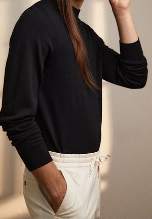Un pull noir à manches longues avec un col montant, associé à un pantalon de jogging crème avec une taille élastique et un cordon de serrage, une poche latérale visible.