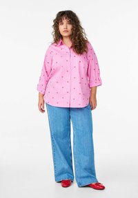 Rosa gestreiftes Hemd mit kleinen roten Herzmustern, mit Kragen, hochgekrempelten Ärmeln, kombiniert mit weit geschnittenen blauen Jeans und roten Schuhen.