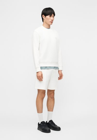 Νεαρός άνδρας που στέκεται φορώντας λευκή φανέλα, λευκά σορτς, λευκές κάλτσες, μαύρα αθλητικά παπούτσια και ανοιχτό μπλε πουκάμισο κάτω από τη φανέλα.